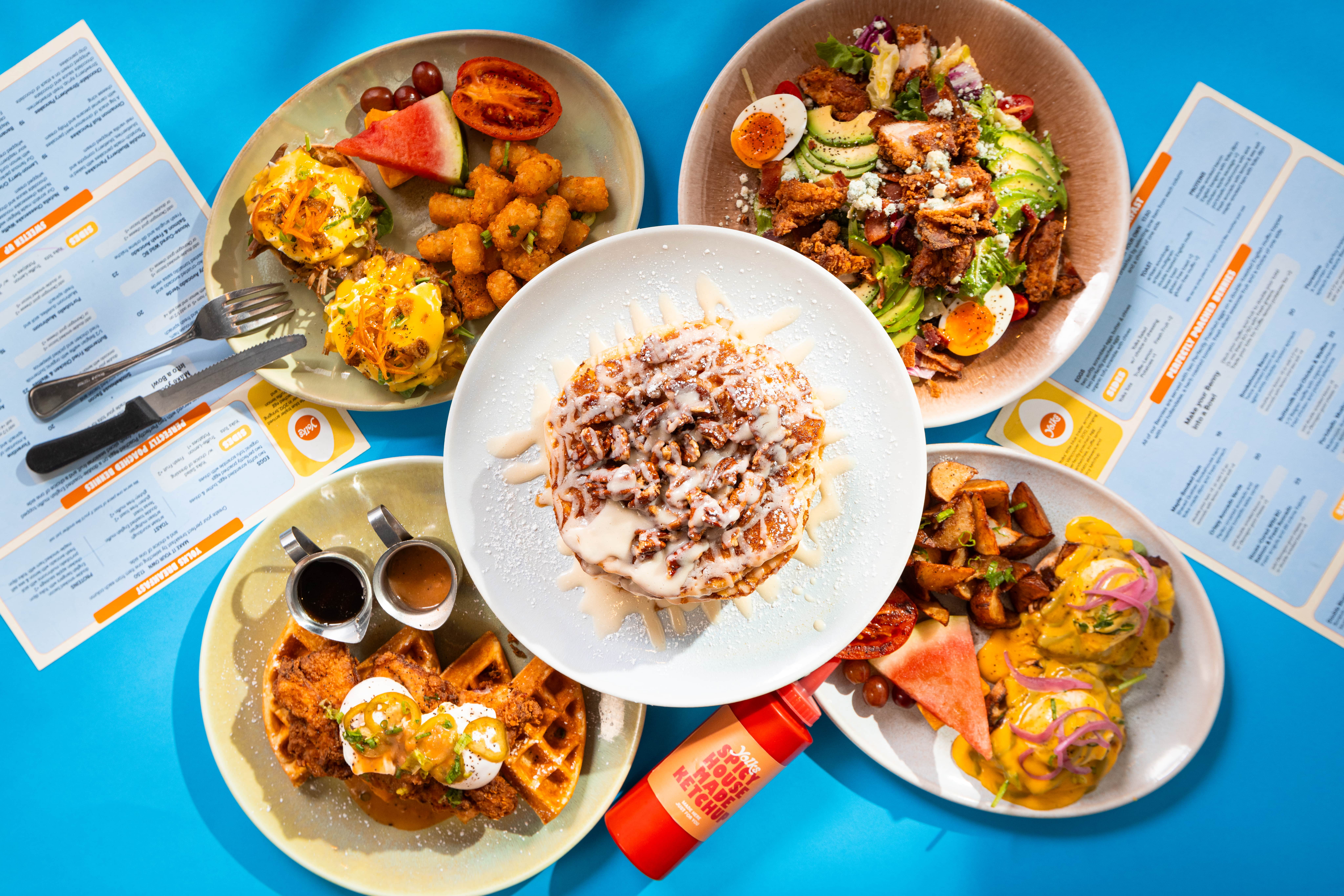 Colorful brunch spread with pancakes, eggs, toast, and breakfast bowls on a blue table