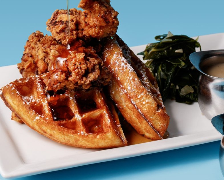 Chicken and waffles breakfast with crispy fried chicken, bacon, and sautéed greens on a plated dish